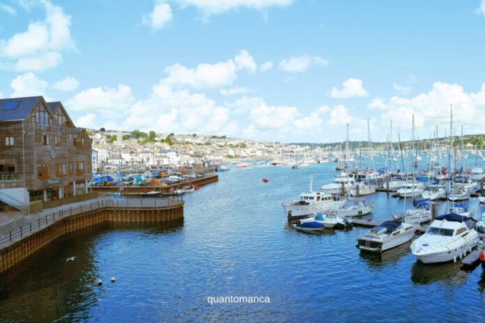 Veduta panoramica del porto storico di Falmouth in Cornovaglia, con numerose barche a vela e imbarcazioni ormeggiate in un'ampia baia naturale sotto un cielo sereno.